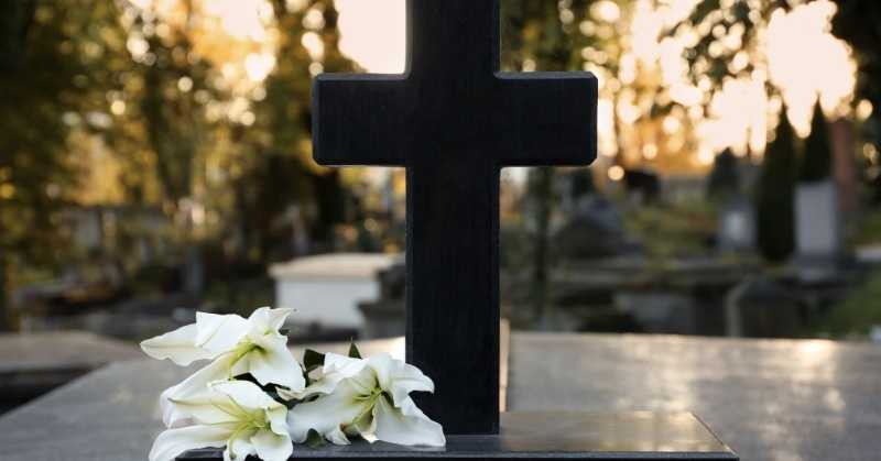 Black cross headstone with white lilies resting at a gravesite in a peaceful cemetery setting.